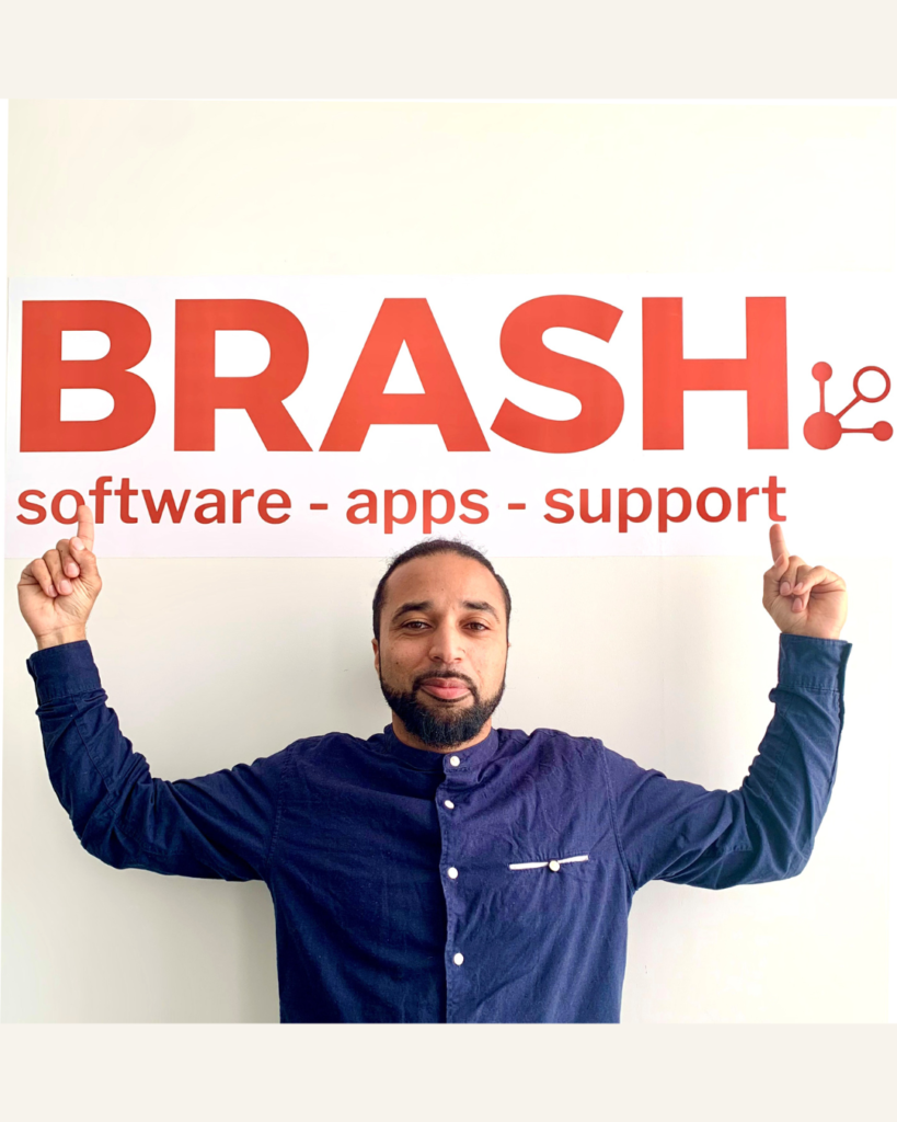 Person standing with arms pointing up wearing a blue button-up shirt against a plain light background and Brash solutions logo in the background.