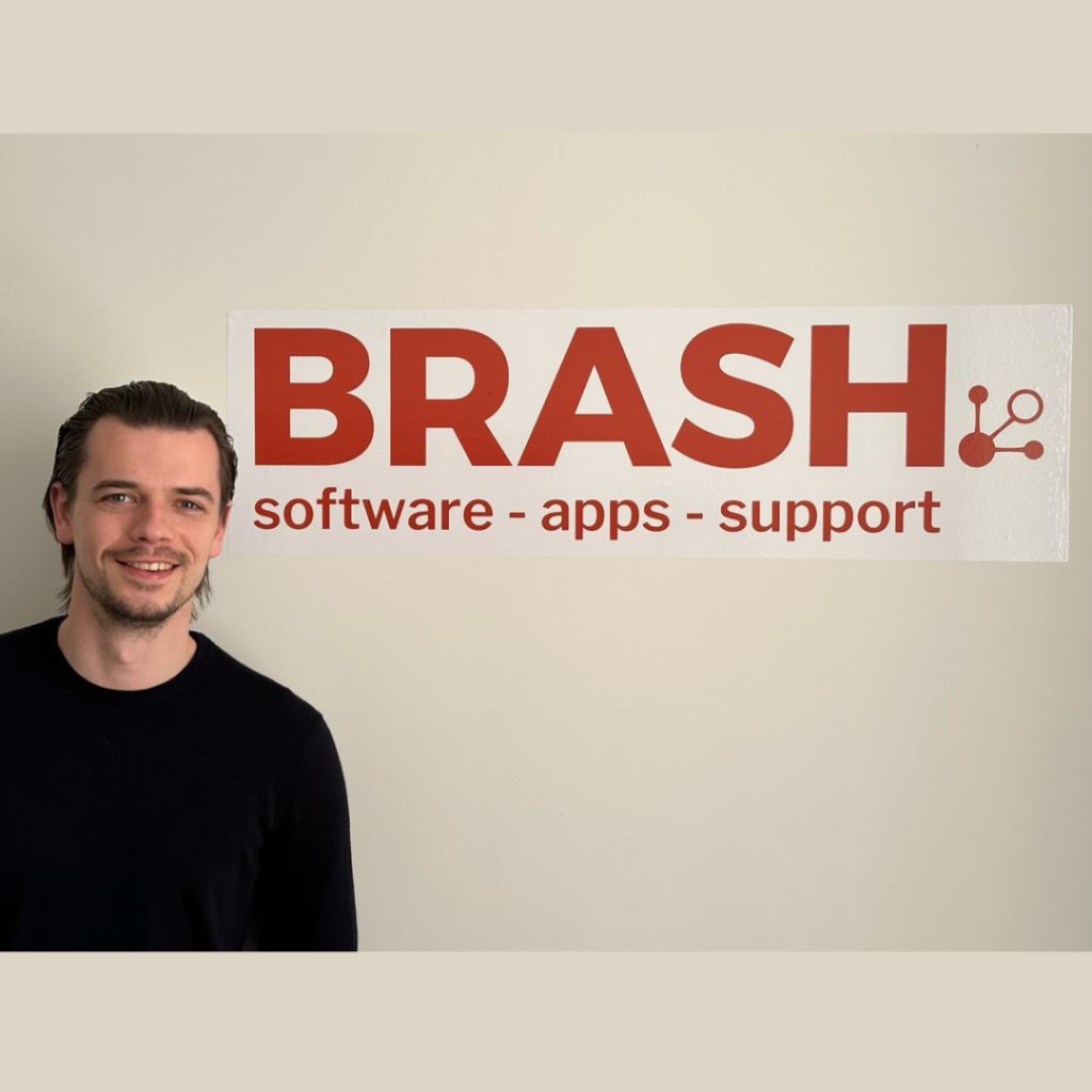 Person standing beside a large Brash Solutions sign displaying the text “BRASH software - apps - support” on a light-colored wall.